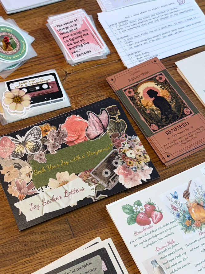 Joy Seeker Letters monthly subscription contents: letter pages, Sacred Word card, music card, recipe card, stickers, laid out on a wood table